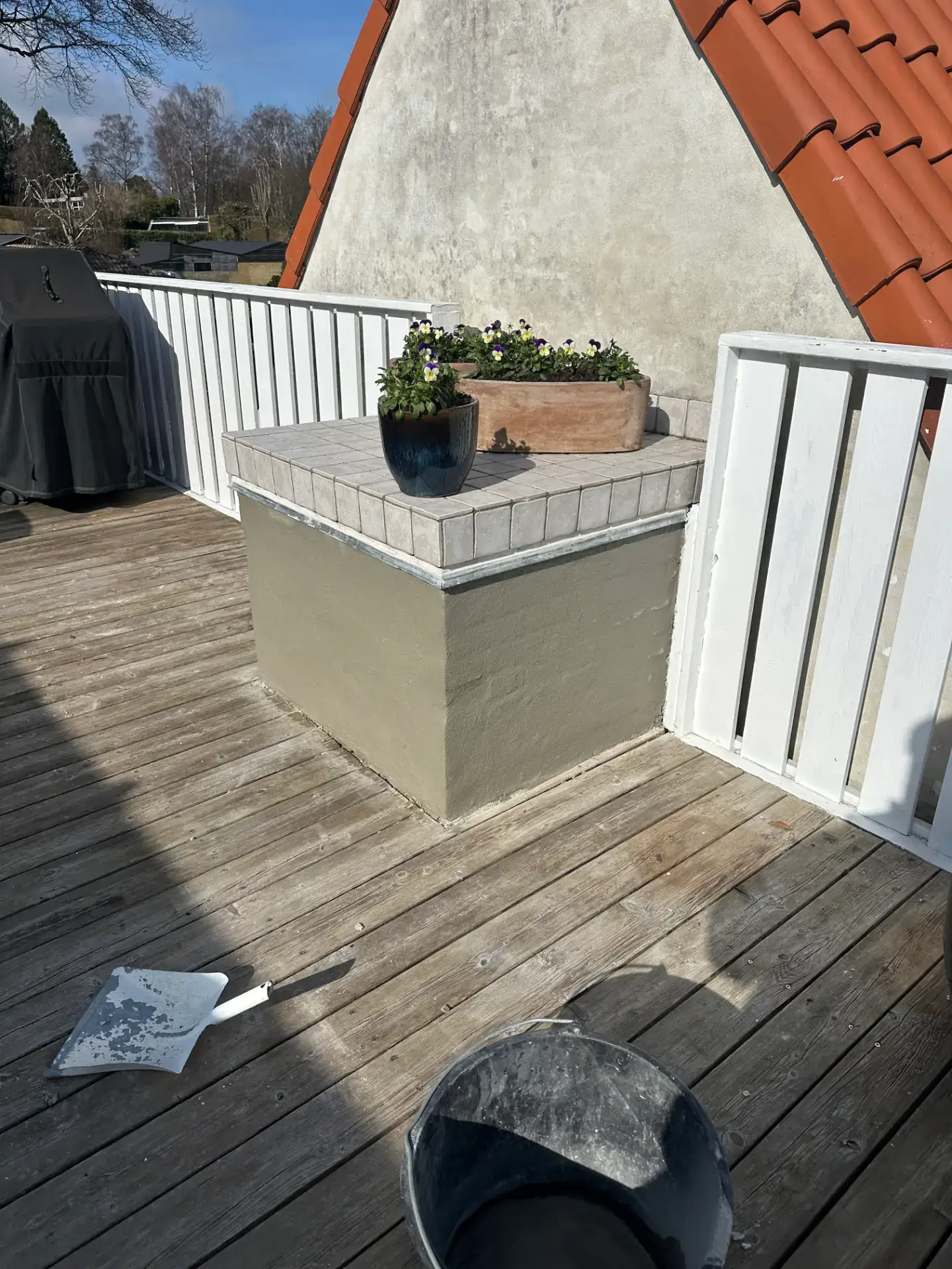 Terrasse med blomsterkasse og udsigt til tag og himmel.